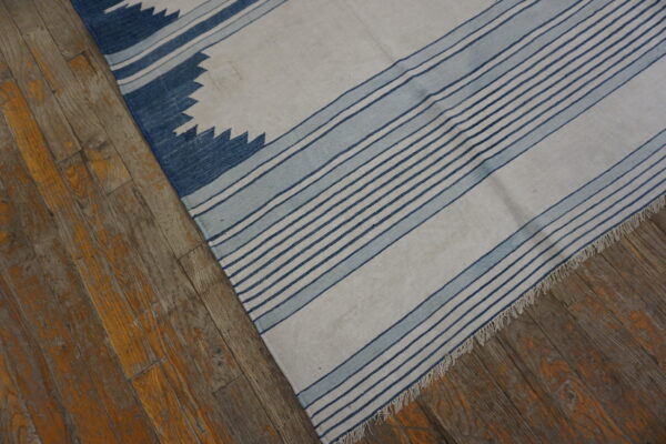 White flatweave rug with blue stripe and sawtooth patterns rests on distressed wooden floorboards.