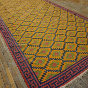 Flatweave rug in gold, navy, red, and green geometric diamonds with a Greek key border on wood planks.
