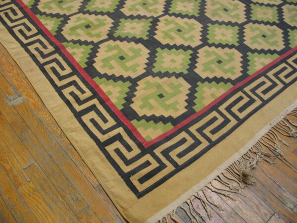 Tan, green, and black flatweave rug featuring a geometric lattice pattern and a greek key border on wood flooring.
