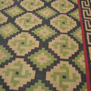 Geometric flatweave rug in tan, green, and black with a red stripe and Greek key border on wood flooring.