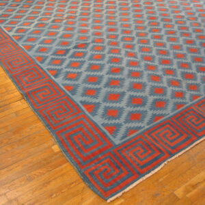 Low-pile blue and reddish-orange rug with a stepped diamond pattern and prominent Greek key border on wood flooring.