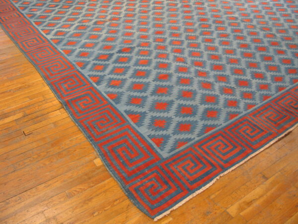 Low-pile blue and reddish-orange rug with a stepped diamond pattern and prominent greek key border on wood flooring.