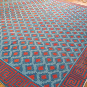 Low-pile blue and orange geometric rug with a thick Greek key border on wood flooring.