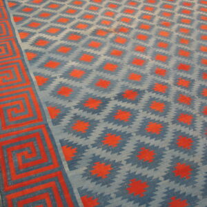 Blue and red flatweave rug with diamond patterns and a Greek key border lies on a wood floor.