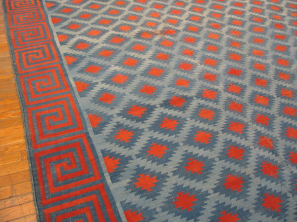Blue and red flatweave rug with diamond patterns and a greek key border lies on a wood floor.