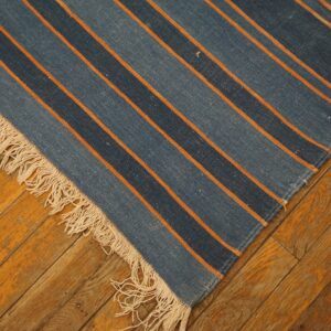 Blue and orange striped flatweave rug with fringe lies diagonally across rustic wooden floorboards.