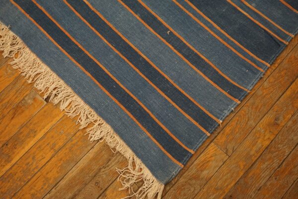 Blue and orange striped flatweave rug with fringe lies diagonally across rustic wooden floorboards.