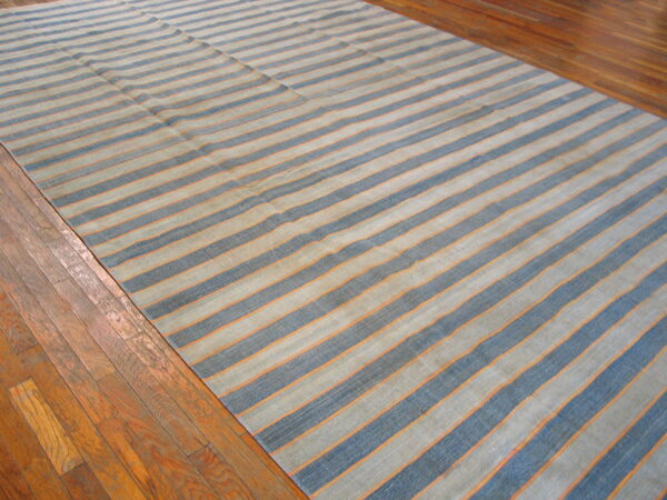 Large, low-pile rug featuring wide blue and pale stripes separated by thin orange lines on hardwood flooring.