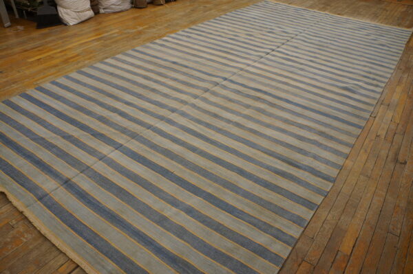 Rectangular flatweave rug featuring blue and beige stripes separated by thin gold lines on a wood floor.