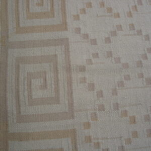 A low-pile beige rug featuring tan concentric squares and square dots rests beside brown wood flooring.