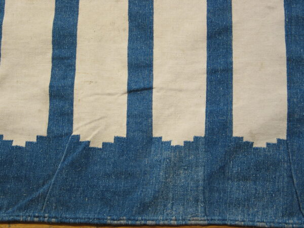 Blue and cream striped flatweave rug showing a stepped edge design resting on a wooden floor.