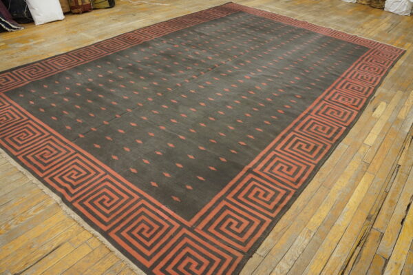 Large dark rug with a red-orange greek key border and dashed pattern on light wood flooring.