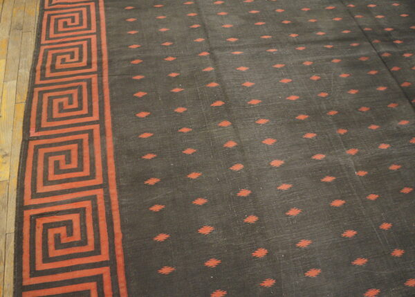 Dark rug featuring a reddish-orange greek key border and small diamond motifs on a light-colored wooden floor.