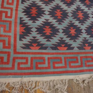 Geometric flatweave rug in red, teal, and navy with a Greek key border resting on wood planks.