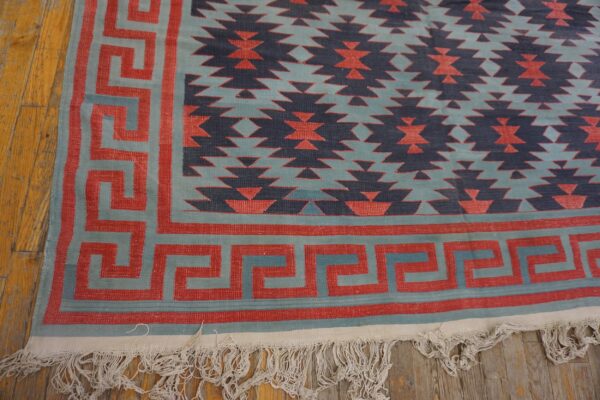Geometric flatweave rug in red, teal, and navy with a greek key border resting on wood planks.