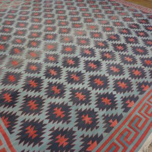 A low-pile geometric rug in teal, navy, and coral features zigzag patterns and a meander border on a wood floor.