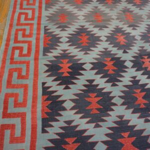 Flat-weave rug with coral Greek key border and navy geometric patterns on distressed wood flooring.