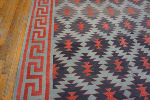 Flat-weave rug with coral greek key border and navy geometric patterns on distressed wood flooring.