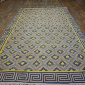 Large blue, yellow, and beige geometric flatweave rug with a Greek key border displayed on worn wood flooring.