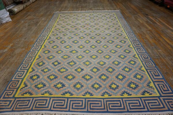Large blue, yellow, and beige geometric flatweave rug with a greek key border displayed on worn wood flooring.