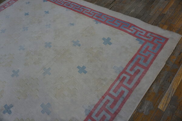 White rug featuring blue plus signs and a pink and blue greek key border, lying on distressed wood flooring.