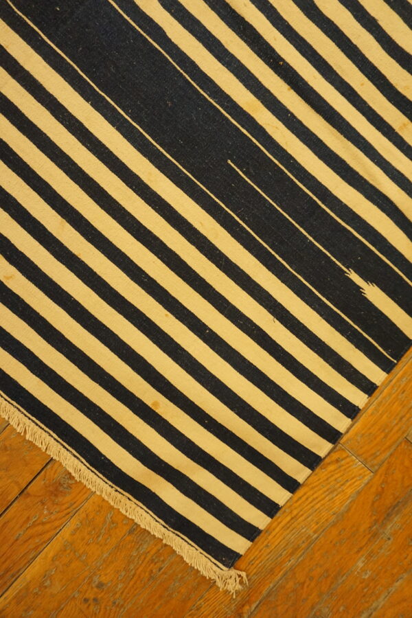 Flatweave rug featuring black and yellow-beige diagonal stripes with fringe, resting on wood flooring.