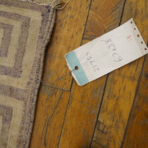 Beige and faded purple flatweave rug with a stepped geometric pattern sits on wood flooring near a tag.