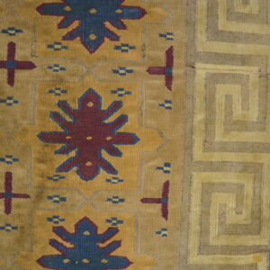 Close-up of a flatweave rug with mustard field, blue and burgundy motifs, and a tan Greek key border on wood floor.
