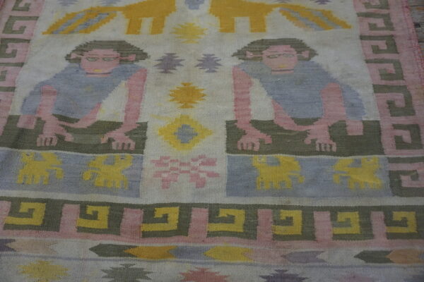 Low-pile rug displaying mirrored human figures and geometric motifs in pink, pale blue, yellow, and olive green.