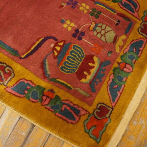 Close-up showing a rosy red rug field and a gold border with multicolored floral and vessel designs on worn wood floors.