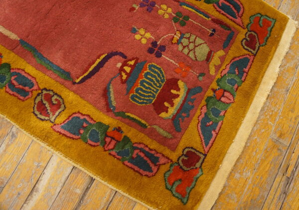 Close-up showing a rosy red rug field and a gold border with multicolored floral and vessel designs on worn wood floors.