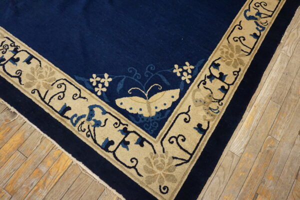 Corner detail of a deep blue rug with a beige floral and butterfly border on weathered hardwood flooring.