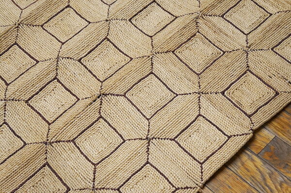 Textured beige rug featuring dark stitching in a geometric tumbling block pattern over rustic hardwood flooring.