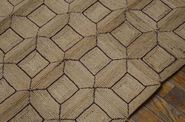 A textured tan rug with dark brown stitching forming an interlocking diamond pattern rests on wood flooring.