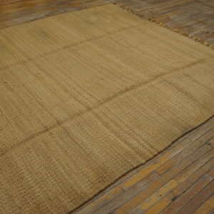 Tan flatweave rug with horizontal seams sits on worn, light brown wood flooring.