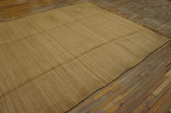Tan flatweave rug with horizontal seams sits on worn, light brown wood flooring.
