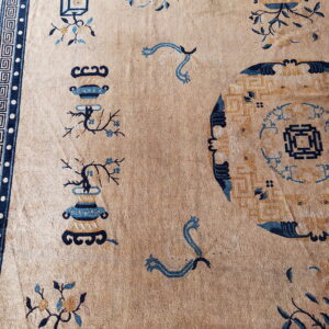 Low-pile tan rug displays blue and ochre vase, floral, and geometric patterns with a thick key border.