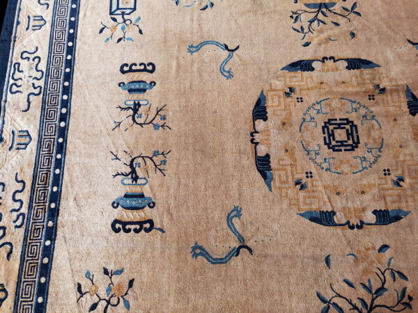 Low-pile tan rug displays blue and ochre vase, floral, and geometric patterns with a thick key border.