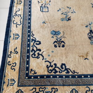 Tan rug featuring a dense dark blue Greek key border and traditional pictorial designs on a white floor.