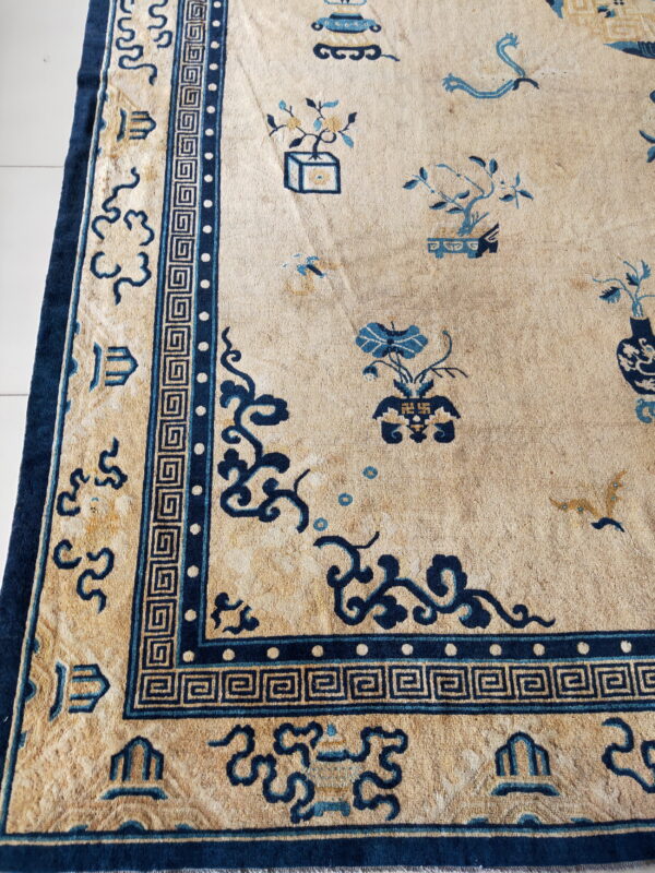 Tan rug featuring a dense dark blue greek key border and traditional pictorial designs on a white floor.