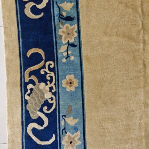 A high-pile tan rug features a complex border strip with dark blue scrollwork and light blue floral patterns.