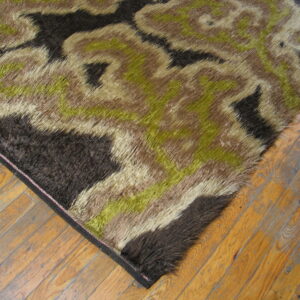 High-pile shag rug in abstract dark brown, olive, and tan patterns rests on distressed hardwood floor.
