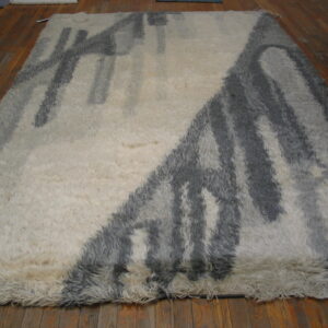 Shaggy, high-pile cream rug featuring a thick abstract grey pattern over reddish-brown wood planks.