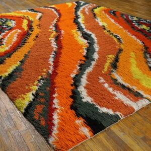 Shaggy pile rug with abstract orange, yellow, green, and black swirls rests on plank wood flooring.