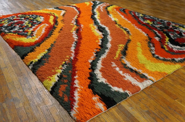 Shaggy pile rug with abstract orange, yellow, green, and black swirls rests on plank wood flooring.