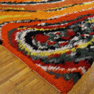 Multi-colored shag rug in red, orange, yellow, and dark green, resting on honey-toned wood floors.