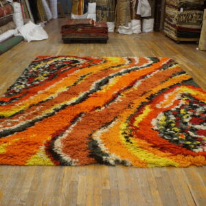Textured high-pile rug with abstract orange, yellow, and multicolored swirling bands on a wooden floor.