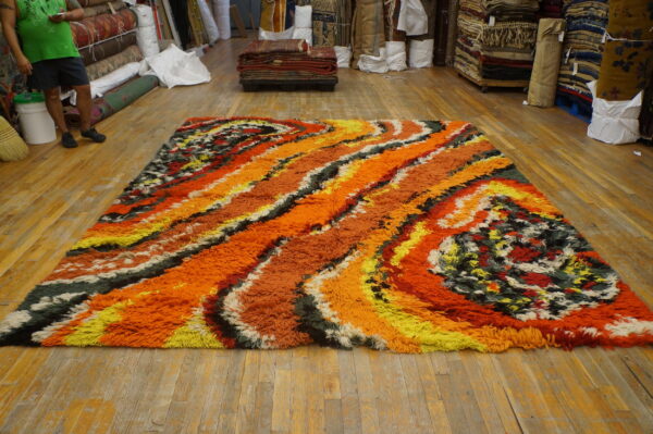 Textured high-pile rug with abstract orange, yellow, and multicolored swirling bands on a wooden floor.