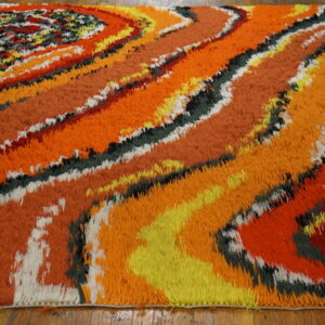 High-pile shag rug in swirling orange, yellow, green, black, and white colors on a wood floor.