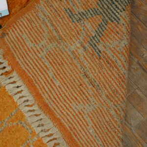 High pile orange rug with gray pattern is folded to show the multicolored striped woven underside and cream fringes.